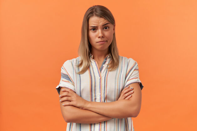 美丽一个穿着条纹衬衫的沉思的年轻女子站着 双臂交叉在橙色的墙上年轻成人姿势