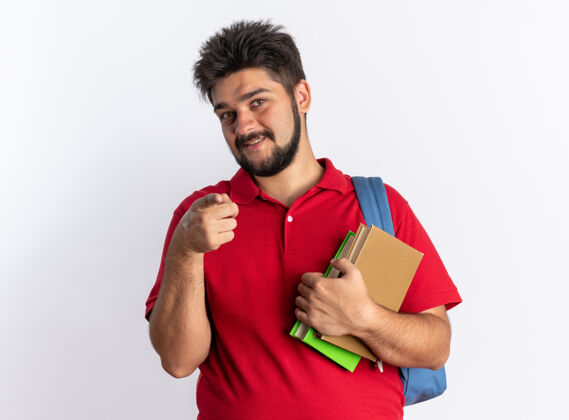 指向年轻的留着胡子的学生 穿着红色马球衫 背着背包 拿着笔记本 用食指指着 站在白色的墙上 快乐而积极地微笑着手势年轻背包