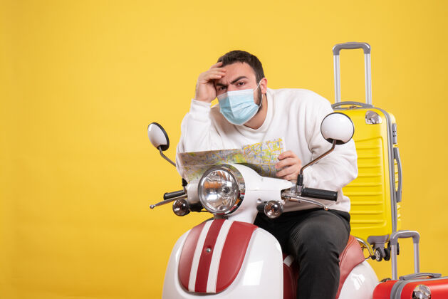 黄色旅行概念和困惑的年轻人在医疗面罩坐在摩托车上 黄色的手提箱上 拿着地图运动车辆人