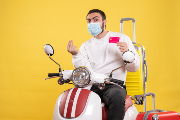面罩旅行概念的正面图 戴着医用面罩的年轻人坐在摩托车上 黄色手提箱 手里拿着银行卡人抱着旅行
