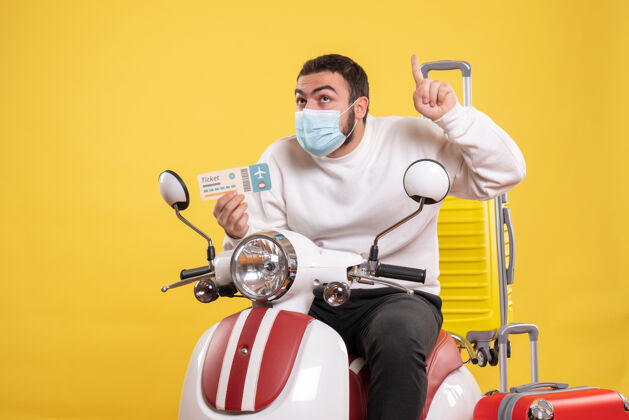 医疗带着医用面罩坐在摩托车上的年轻好奇的家伙的旅行概念的顶视图票手提箱举着