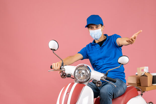 摩托车前视图好奇的快递员戴着医用面具戴着帽子坐在滑板车上传递粉彩桃色背景的命令背景穿医疗