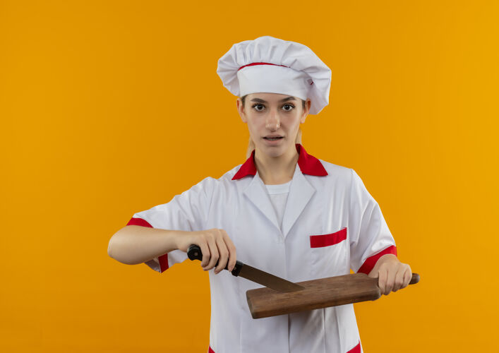 切割年轻漂亮的厨师穿着厨师制服拿着刀和砧板孤立在橙色的墙上厨师持有制服