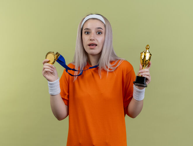 奖杯惊讶的年轻白人运动女孩戴着背带 戴着头带和腕带 拿着金牌和冠军杯腕带年轻冠军