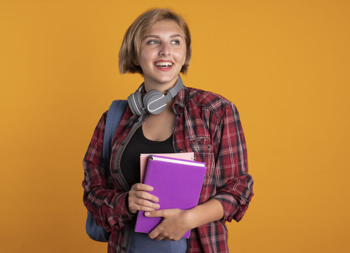 微笑微笑的年轻斯拉夫女学生脖子上戴着耳机 背着书包 手里拿着书和笔记本 看着身边拿着周围穿