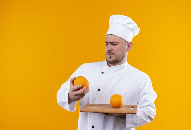 烹饪迷茫的年轻帅哥厨师身着厨师制服拿着砧板和橘子 看着隔离在橘子墙上的它切着板子厨师