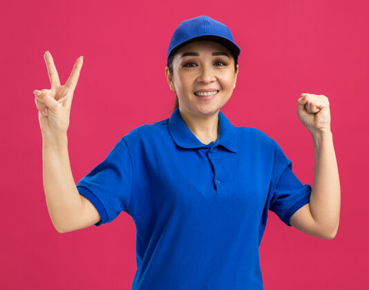 握紧身穿蓝色制服 头戴鸭舌帽的年轻女送货员站在粉红色的墙上高兴地微笑着 手里握着v字拳头帽子制服传递