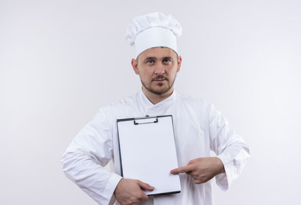 帅气自信的年轻帅哥厨师 身着厨师制服 拿着并指着隔离在白墙上的剪贴板年轻制服厨师