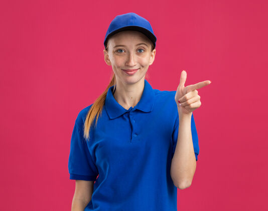 微笑身穿蓝色制服 头戴鸭舌帽的年轻送货员友好地用食指指着站在粉色墙上的一侧制服指点朋友