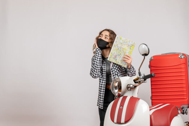 站立戴着面具拿着地图的年轻漂亮女孩站在带着手提箱的轻便摩托车旁边人物手提箱黑色