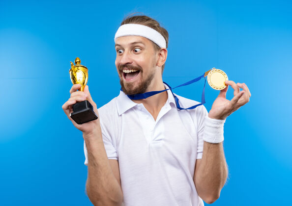 奖牌快乐的年轻英俊的运动型男子戴着头带和腕带 脖子上戴着奖牌 手里拿着奖牌和优胜者杯 看着隔离在蓝色墙上的奖杯运动男人穿