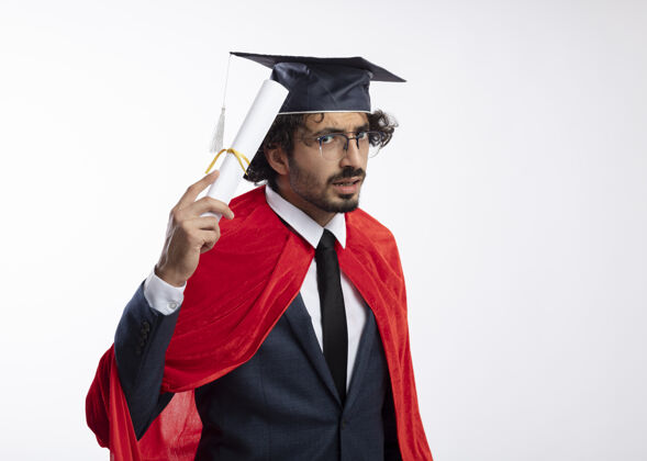 斗篷无知的年轻白种人超级英雄戴着眼镜 穿着红色斗篷的西装 戴着毕业帽 手里拿着毕业证书眼镜不知所措帽子