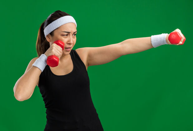 健身戴着头带和哑铃做运动的年轻健身女士紧张而自信地站在绿色的墙上年轻头带紧张