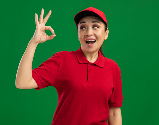 靠边站身穿红色制服 头戴帽子的年轻女送货员站在绿色的墙上 高兴地看着旁边的“一切正常”标志接生好的帽子