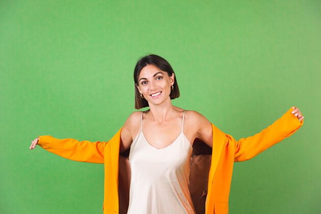 成人穿着丝绸米色连衣裙和橙色超大西装的时尚女士 身着绿色 带着积极的情绪微笑积极站立女性化