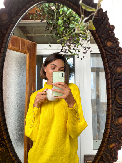 公寓家里的美女穿着舒适温暖的黄色毛衣 用手机在镜子里自拍故事和在社交媒体上发帖房子自我旅游