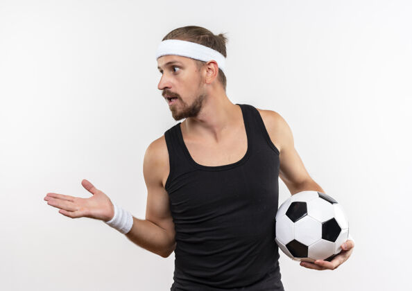 年轻令人印象深刻的年轻英俊的运动男子戴着头带和手镯拿着足球看一边显示空手隔离在白色的墙壁上手足球英俊