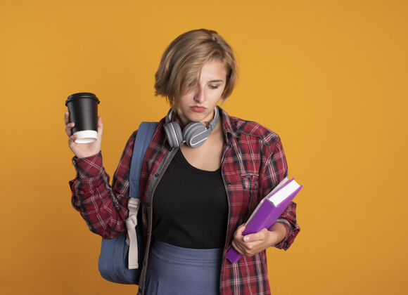 困惑困惑的年轻斯拉夫学生女孩戴着耳机 背着背包 拿着纸杯 看着书和笔记本学生空间笔记本