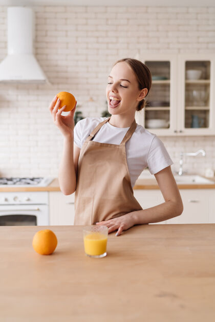 桌子年轻漂亮的女人在厨房里围着围裙 水果和橙汁护理肖像素食