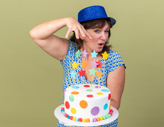 举行中年妇女戴着派对帽 手拿生日蛋糕 微笑着站在绿色的墙上欢庆生日派对欢呼生日女人