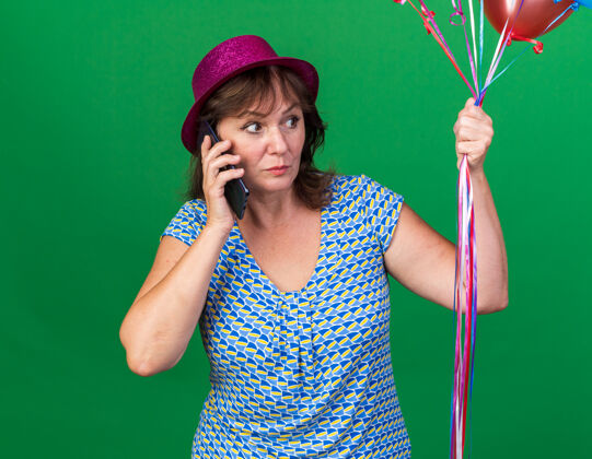 立场中年妇女戴着派对帽 手里拿着五颜六色的气球 一边说着手机 一边站在绿色的墙上庆祝生日派对庆祝女人而