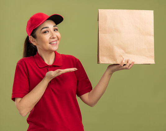 介绍快乐的年轻送货员 身穿红色制服 戴着帽子 手里拿着纸包 脸上带着微笑 手举着胳膊站在绿色的墙上帽子手举行