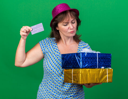 帽子中年妇女戴着派对帽 手里拿着生日礼物和信用卡 神情困惑地站在绿色的墙上庆祝生日派对立场困惑女人