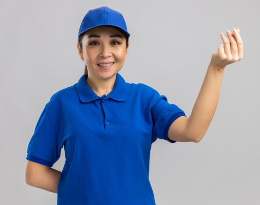 年轻身穿蓝色制服 戴着帽子的年轻送货员站在白色的墙上做着搓手指的赚钱手势 开心地微笑着手指钱制服