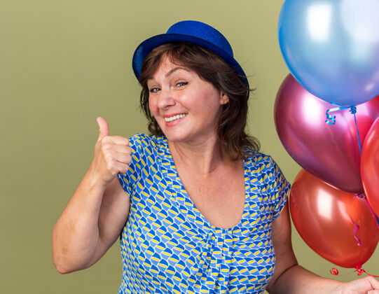 五颜六色快乐的中年妇女戴着派对帽 手里拿着一堆五颜六色的气球 微笑着竖起大拇指 站在绿色的墙上庆祝生日派对微笑年龄生日