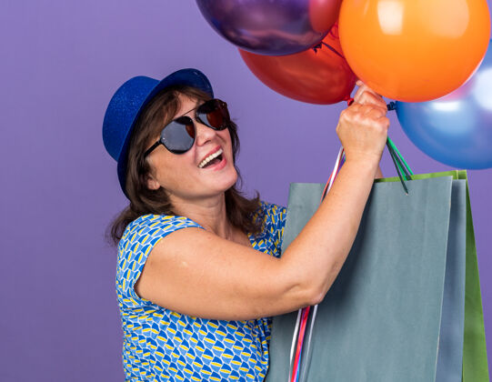 立场快乐快乐的中年妇女戴着派对帽戴着眼镜拿着一束五颜六色的气球和带着礼物的纸袋微笑着站在紫色的墙上庆祝生日派对女人包束