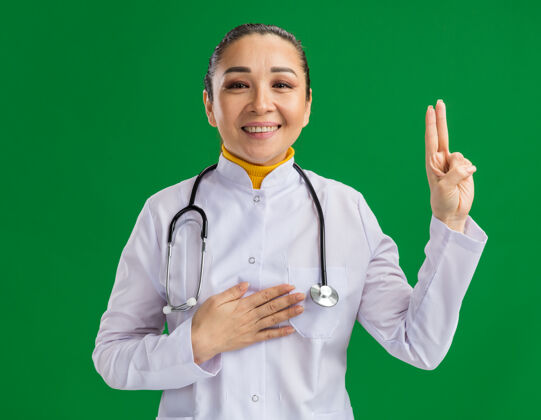 女人快乐的年轻女医生 穿着白药衣 脖子上戴着听诊器 手放在胸前宣誓 手指站在绿色的墙上胸部发誓手指