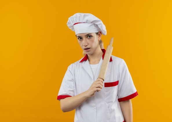 年轻年轻漂亮的厨师穿着厨师制服 拿着擀面杖 站在橙色的墙上 留着复制空间漂亮制服烹饪