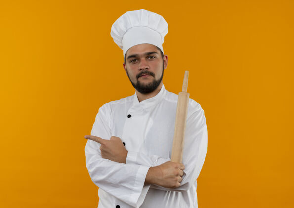 制服自信的年轻男性厨师 身着厨师制服 站着 姿势封闭 拿着擀面杖 指着橙色墙壁上的隔离墙 留有复制空间年轻姿态厨师