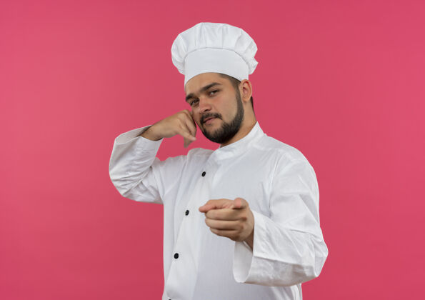 呼叫自信的年轻男厨师身着厨师制服 在粉色墙壁上做着呼叫手势 并用复制空间孤立地指指点点厨师烹饪年轻