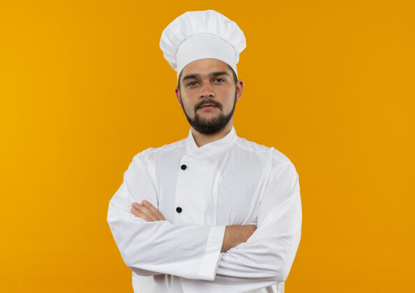 烹饪自信的年轻男性厨师身着厨师制服 以封闭的姿势站在橙色墙壁上 并留有复制空间自信年轻封闭