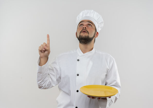 空的穿着厨师制服的年轻男厨师手拿空盘子 孤零零地站在白色的墙上看着 指着上面制服盘子厨师
