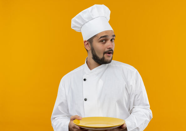 盘子令人印象深刻的年轻男厨师在厨师制服举行空盘子隔离在橙色墙上空制服烹饪
