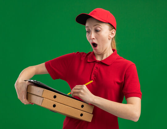 铅笔身穿红色制服 头戴鸭舌帽 拿着披萨盒 夹着空白纸的剪贴板 拿着铅笔的年轻送货女孩 看上去很惊讶地站在绿色的墙上写着什么站着写字披萨