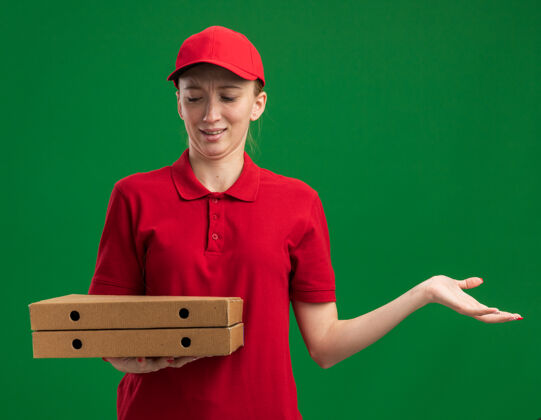 年轻穿着红色制服 戴着帽子 拿着披萨盒的年轻送货女孩站在绿色的墙上 困惑地看着披萨盒拿着女孩制服