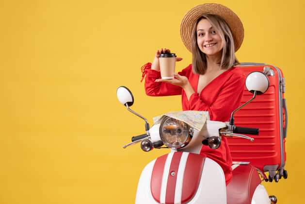 人微笑着的穿着红裙子的年轻女士拿着咖啡杯靠近轻便摩托车时尚杯子微笑的年轻女士