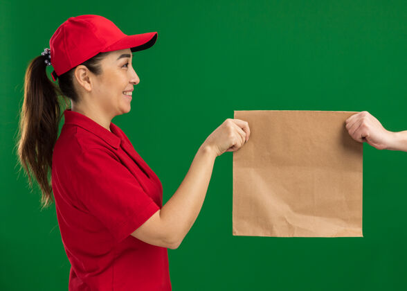 送货年轻的送货员身着红色制服 头戴鸭舌帽 站在绿色的墙边 微笑着自信地给顾客送纸包给予微笑包装