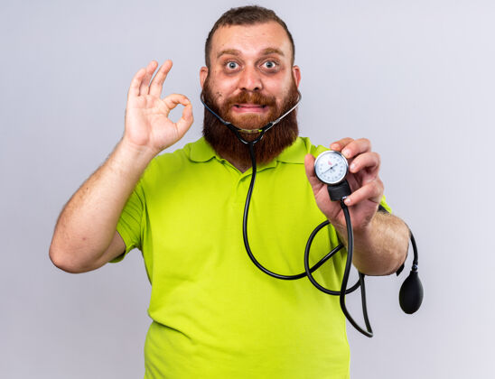 胡须不健康的胡子男人穿着黄色马球衫感觉不舒服用血压计测量血压困惑显示ok标志站在白色的墙上站压力感觉
