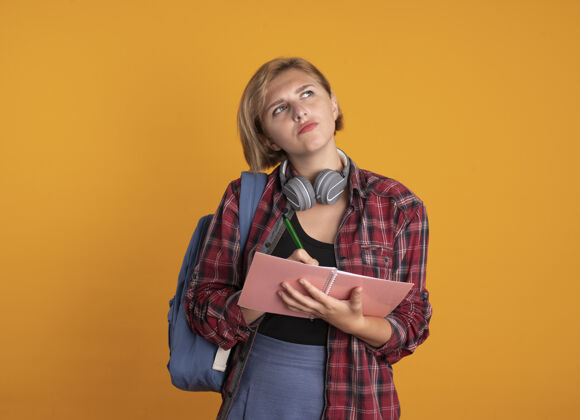 拿着体贴的年轻斯拉夫女学生戴着耳机 背着背包 拿着笔记本和笔 抬头望去斯拉夫人笔记本背包