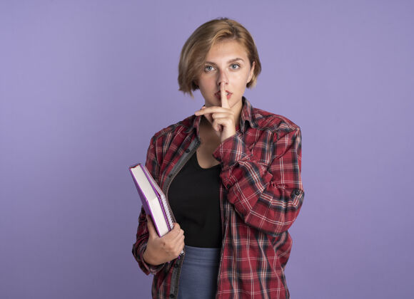 斯拉夫自信的年轻斯拉夫学生女孩拿着书和笔记本手势沉默的标志在紫色背景上与复制空间隔离笔记本紫色标志