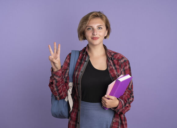 拿着高兴的年轻斯拉夫学生女孩背着书包拿着书和笔记本用三个手指做手势复制三手势