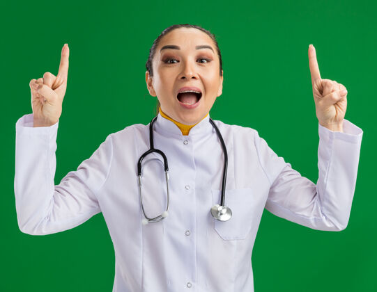 年轻人身穿白色药衣 脖子上戴着听诊器的年轻女医生高兴又惊讶地向站在绿墙上的有新想法的指导员们展示了自己的想法Figner医生听诊器