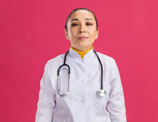 听诊器身穿白色药衣 脖子上戴着听诊器的年轻女医生站在粉红色的墙上 表情自信外套站表情