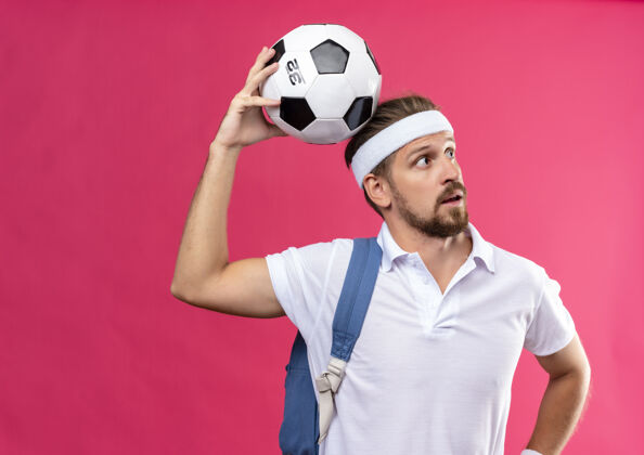 穿令人印象深刻的年轻英俊的运动型男子戴着头带和手镯 肩上背着书包 看着侧面 头上举着足球隔离在粉红色的墙上头头带腕带