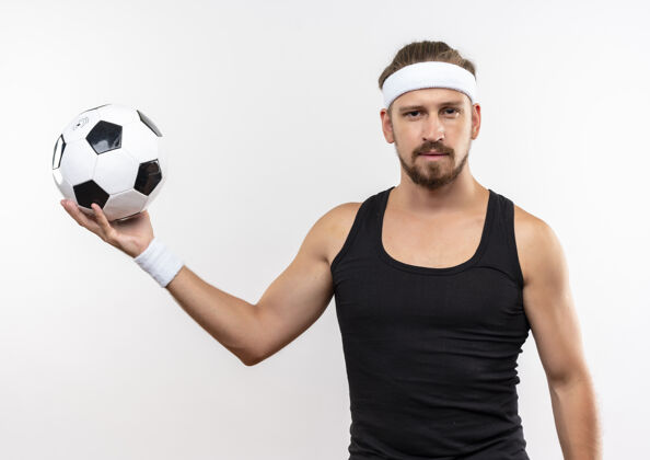 年轻自信的年轻帅气的运动型男人戴着头带和腕带拿着足球 在白墙上显得孤立无援腕带男人足球
