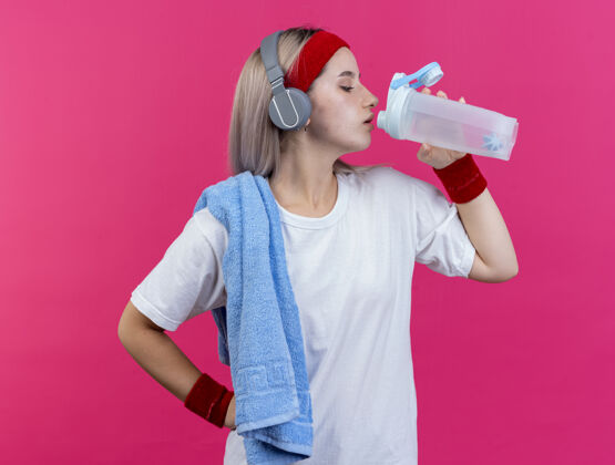 肩膀自信的年轻白人运动女孩戴着耳机戴着背带戴着头带和腕带肩上拿着毛巾从水瓶里喝水复制耳机运动
